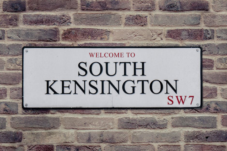 A rectangular white sign mounted on a brick wall displaying the text 'Welcome to South Kensington SW7' in black and red lettering. The brick wall behind the sign consists of reddish-brown bricks with visible mortar joints. The sign is positioned at eye level, suggesting an outdoor setting near a property's entrance, which is relevant for home relocation or moving services. This image captures the signage often encountered in residential or urban areas of South Kensington, a common destination during furniture transport and packing and moving processes. The clean, professional appearance of the sign and brick backdrop reflect a typical setting where removals or property changes are organized and managed, as seen in professional relocations by companies such as Man and Van Kensington.