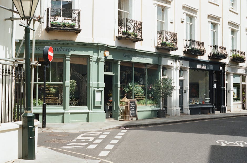 A street scene in Kensington showing a three-story building with a light green ground-floor shopfront and several upper-floor residential windows with black wrought iron balconies. The shop appears to be a florist or flower shop, with large glass windows displaying plants and flowers, and a small blackboard sign placed on the sidewalk outside. On the pavement, a curbside area has a zebra crossing with painted white dashed lines leading to the shop entrance. Adjacent to the shop, there is a black van partially visible, with a scene of moving activity, including stacked cardboard boxes, moving blankets, and packing materials being loaded or unloaded. Visible tools include a trolley, and the loading process is taking place on the street in front of the property, illustrating logistics related to home relocation or furniture transport. The environment features soft natural lighting, and the overall scene captures elements typical of residential street moving services, with Man and Van Kensington specialising in removals.