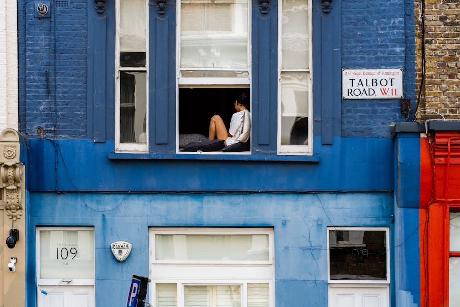 A woman sitting on an open window ledge of a blue-painted residential building on Talbot Road, Kensington, observing her surroundings during daylight. The window is wide open, revealing her seated with legs crossed, wearing a white shirt and black shorts. Inside, a bed with a grey headboard and white bedding is partially visible behind her. The building’s facade features decorative wooden trim and multiple small windows, with some open and others closed, alongside a street sign indicating 'Talbot Road, W.11.' Below, the ground-floor entrance door displays the number '109' and a security alarm system. The area appears to be a typical urban residential setting with neighboring buildings painted in similar bright colors, and a parking sign visible near the door, hinting at local parking regulations. This scene subtly connects to house removals and home relocation services, highlighting aspects of moving logistics such as staging, building access, and the typical environment encountered during packing and furniture transport in central London.