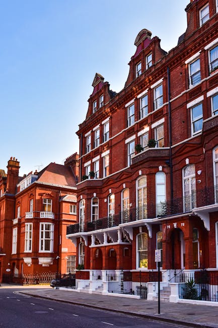 The image depicts a row of historic red-brick residential buildings with large windows and decorative iron balconies, situated along a quiet street on a clear day with blue skies. In the foreground, a moving van with its rear door open is parked close to the pavement, revealing a stacking of cardboard boxes, wrapped furniture, and moving blankets inside. A hand truck or trolley with a covered piece of furniture is positioned nearby, mid-movement, indicating an active loading process. The scene suggests a home relocation or furniture transport scenario, supported by the presence of packing materials, protective coverings on large items, and the vehicle ready for moving logistics. Man and Van Kensington, a professional removals service, is likely handling this household move, and the setting emphasizes careful furniture handling and packing considerations typical of house removals in Kensington, London.