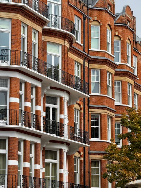 The image shows a row of multi-story Victorian-style terraced houses on a busy street in Kensington, London, with detailed cream-colored exteriors, large windows, and decorative cornices. In the foreground, pedestrians and cyclists are crossing the road, including a man on a green bicycle, a person walking with a backpack, and another cyclist. Parked cars line both sides of the street, with a mix of small hatchbacks and larger vehicles. The sky is blue with scattered clouds, and some buildings have scaffolding, indicating ongoing construction or renovation. The scene captures an active residential area typical of house removal and home relocation settings. The atmosphere suggests daytime activity, suitable for professionals such as those from Man and Van Kensington, specialising in removals and furniture transport, ensuring smooth packing and moving logistics.