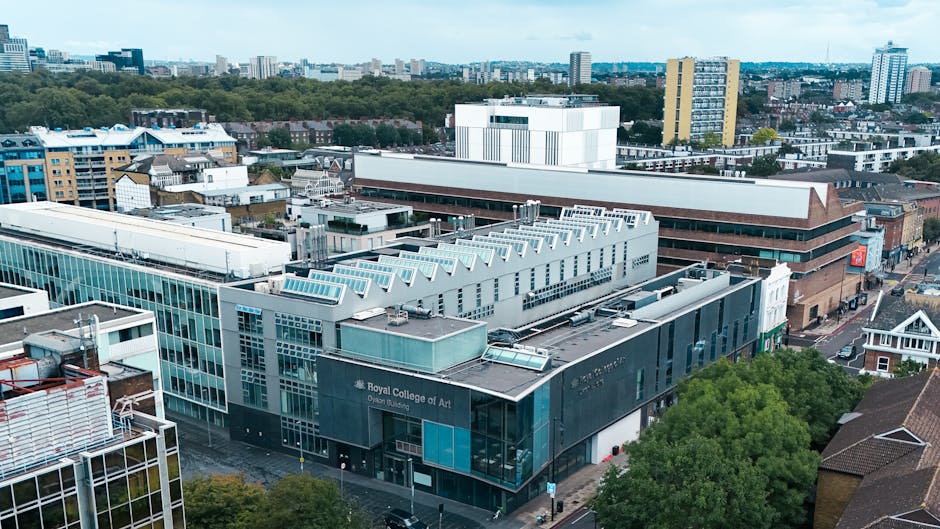 An aerial view of a cityscape showing the exterior of the Royal College of Art building in the foreground, with modern glass and concrete architecture, adjacent to other commercial and residential buildings. The scene includes rooftop equipment such as vents and solar panels. In the background, there are trees, low-rise buildings, and taller high-rise structures under a partly cloudy sky. The image depicts an urban environment suitable for house removals and furniture transport, capturing the setting where professional movers like Man and Van Kensington might operate during home relocation or packing and moving activities within the area.