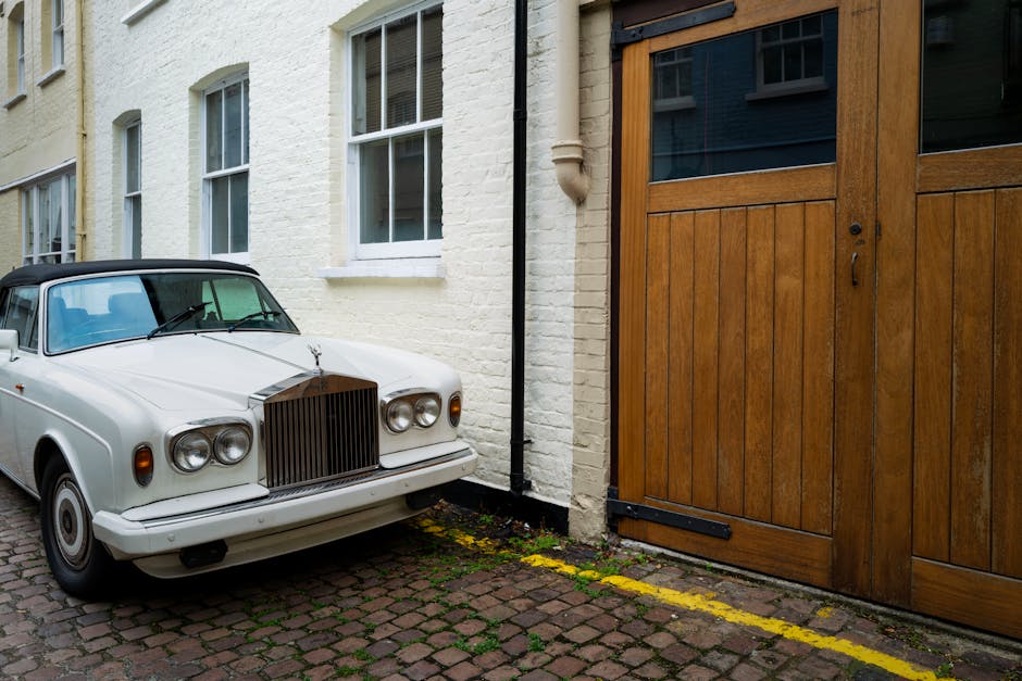 A vintage white car with a black convertible roof parked on a cobblestone driveway beside a white-painted brick house with three visible windows. Adjacent to the vehicle is a large wooden sliding gate with glass panels at the top, positioned next to the house. The driveway features a yellow parking boundary line near the gate, indicating designated parking space, while the pavement shows moss and dirt accumulation. Inside the property, a partially visible interior area suggests the presence of household furniture or moving boxes, although not clearly visible in this outdoor setting. The scene is well-lit with natural daylight, underscoring the structural details of the house, vehicle, and driveway, relevant to home relocation and furniture transport considerations. Man and Van Kensington’s removal services often include handling such objects during loading and unloading processes, respecting parking rules within the Royal Borough of Kensington and Chelsea.