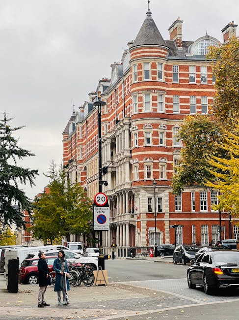 A busy street scene in Kensington featuring a large, multi-storey red-brick building with white stone accents, turrets, and decorative windows. In the foreground, two men are engaged in conversation near parked bicycles and a small wooden sign, with one of them wearing a blue jacket and the other in dark clothing. Several cars are parked along the street and a black vehicle is seen in motion at the intersection. A streetlight and a speed limit signage indicating 20 mph are visible, with trees showing autumnal foliage on the right side of the image. The scene depicts urban residential or commercial architecture typical of a home relocation area, highlighting the context of house removals, relocation services, and move logistics, consistent with the services offered by Man and Van Kensington.