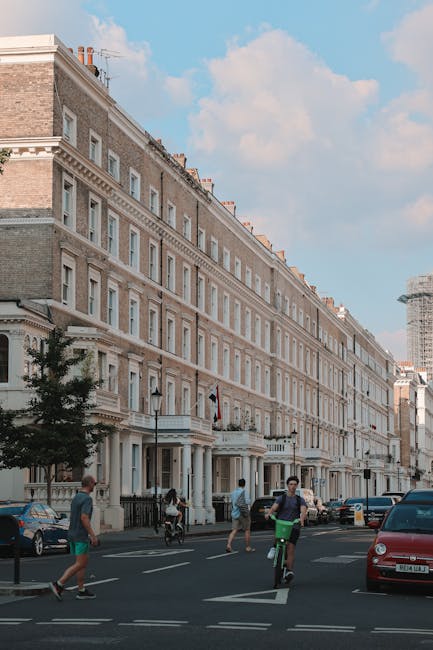 Photograph of a typical London street scene in South Kensington, showing a row of elegant Victorian-style terraced houses with cream and beige brick facades, white window frames, and black wrought iron railings. The buildings are several stories high with chimney stacks on the roofline, and some have flags displayed on the front. In the foreground, a wide pavement features pedestrians walking, including a man in a grey T-shirt and shorts, another person pushing a bicycle, and a woman in casual attire. A black parked car and a red vehicle are visible on the street, along with a few other cars parked along the roadside. The scene is well-lit with natural daylight, under a partly cloudy sky, suggesting daytime conditions. This image captures elements relevant to house removals or home relocation services, such as the urban setting, residential building architecture, and the presence of residents and vehicles involved in everyday activities, reflecting the context of furniture transport and moving logistics associated with South Kensington house removals by Man and Van Kensington.