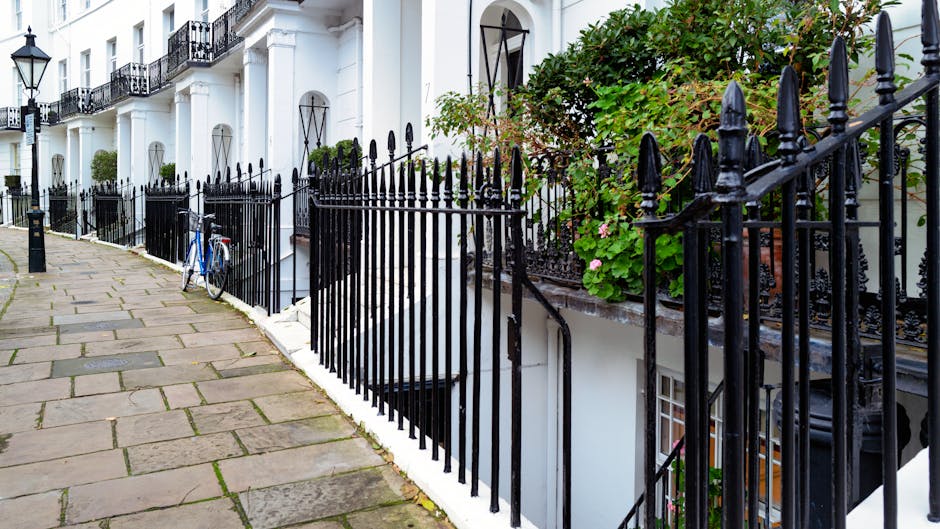 A row of white terraced houses with black wrought-iron fencing along the pavement, which features large stone slabs, in a residential area of South Kensington. A bicycle is secured to the fence near the entrance of one house. Green foliage and pink flowers grow along the fence and on the balconies, which have black decorative railings. The scene appears to be in daylight, capturing a calm street environment. In the context of house removals or furniture transport, this setting reflects the exterior environment where unloading or loading may occur, with the fencing and pavement providing access points for a professional moving team, potentially involving the use of removal vehicles or trolleys outside the property, as part of a home relocation process. Man and Van Kensington operates in this area, offering professional removals and moving services for properties like these.
