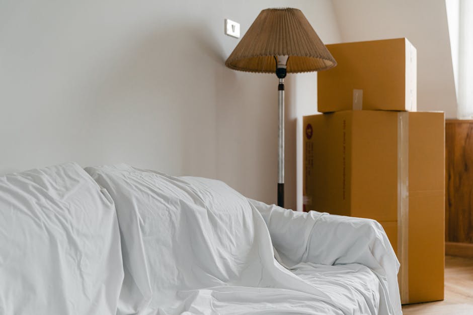 Image showing a room with a white sofa covered in a protective white sheet, positioned next to a wall with a wall socket visible. To the right, there are three large cardboard boxes stacked, with the smallest on top, all secured with packing tape. A tall floor lamp with a beige lampshade is situated behind the boxes, next to a wooden cabinet and a window with sheer curtains allowing natural light into the space. The scene captures elements of house relocation, including packing and furniture moving, with the boxes ready for transportation. The environment appears tidy, and the objects are arranged for easy loading onto a removal van, illustrating a typical interior during a home move or packing process. Man and Van Kensington typically handles such furniture transport and moving logistics, as seen in this staging area for removals.