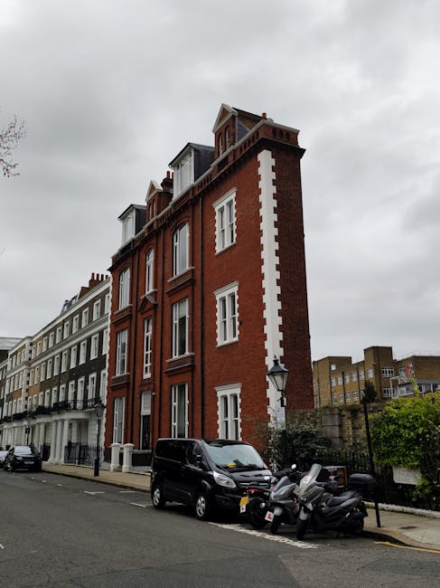 The image shows the exterior of a multi-storey red brick building located on a residential street near Gloucester Road in Kensington, London. The building features white window frames and decorative architectural details, with a small rooftop structure visible at the top. Adjacent buildings on the left are painted in lighter colours, and the street is lined with parked cars and scooters, including a black van parked directly in front of the building. The pavement appears clean, and there is a lamppost visible next to the parked vehicles. Overcast grey skies cast diffuse lighting over the scene, typical of urban residential areas. This setting is relevant to house removals and furniture transport services, such as those provided by Man and Van Kensington, illustrating the type of urban environment where home relocation and moving logistics might take place.